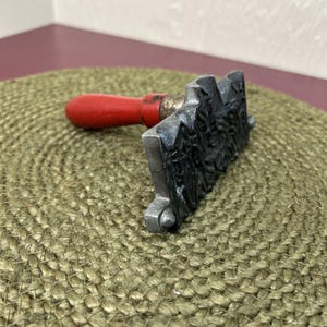 May include: A vintage metal stamp with a detailed design. It features a red wooden handle and a rectangular stamping surface. The stamp is resting on a woven, circular mat.