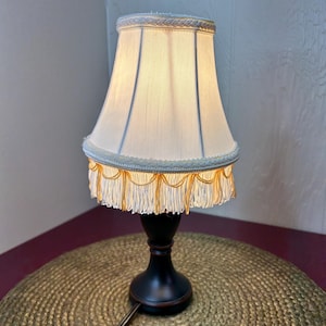 May include: A small, decorative table lamp with a cream-colored lampshade. The shade is trimmed with a light blue and gold fringe. The lamp has a black base and a power cord.