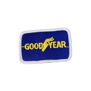 May include: A rectangular patch with a blue background and the yellow text "GOOD YEAR". The patch has a white border and features a yellow winged foot logo, commonly associated with the Goodyear tire brand.