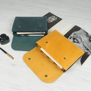 May include: Two leather journals in teal and mustard yellow are displayed open and closed on a light wood surface. Each journal has a flap closure with a brass button. An ink bottle and pen are also visible.