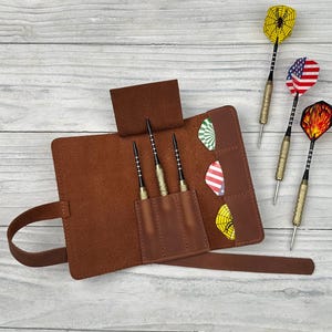 Peut inclure: Un étui à fléchettes en cuir marron avec trois fléchettes à l'intérieur et trois fléchettes avec des plumes décoratives. L'étui est ouvert, révélant les fléchettes et les poches. Les plumes ont divers motifs, dont une toile d'araignée, le drapeau américain et des flammes.