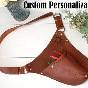 May include: A brown leather tool belt with a pocket and a strap. The belt has a metal ring and is personalized with the text "HOOKS & HAVE".
