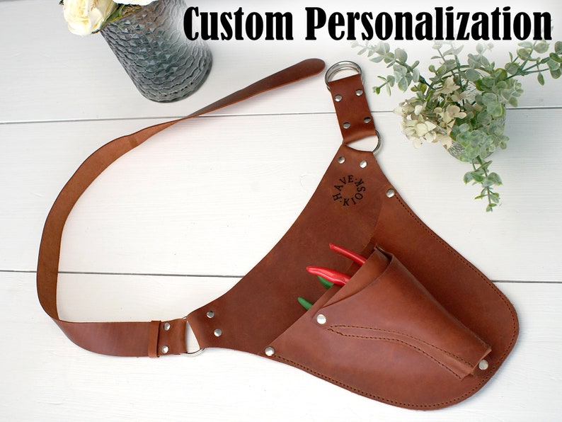 May include: A brown leather tool belt with a pocket and a strap. The belt has a metal ring and is personalized with the text "HOOKS & HAVE".
