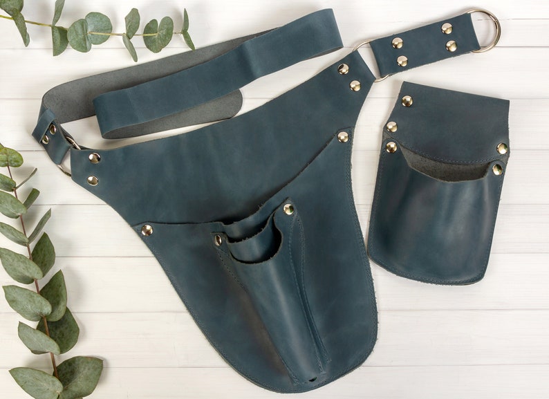 May include: A blue leather tool belt with a large pocket and a smaller pocket on the side. The belt has a metal buckle and is secured with metal rivets.