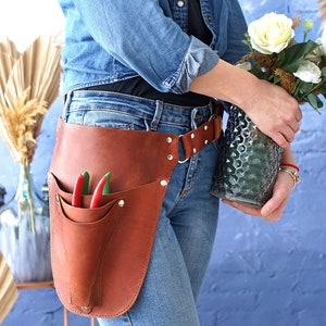 May include: A brown leather gardening tool belt with a pocket for tools and a belt loop. The belt is worn by a person holding a vase of white flowers.