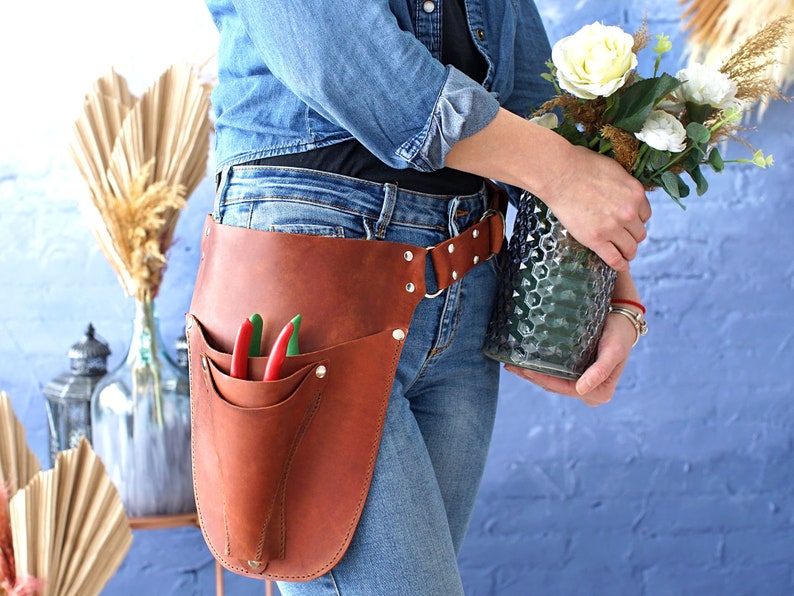 May include: A brown leather gardening tool belt with a pocket for tools and a belt loop. The belt is worn by a person holding a vase of white flowers.