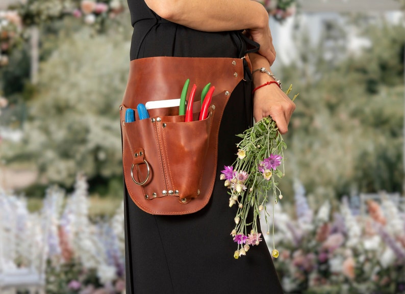 May include: A brown leather tool belt with multiple pockets and a metal ring. The belt is worn by a person holding a bouquet of purple flowers.