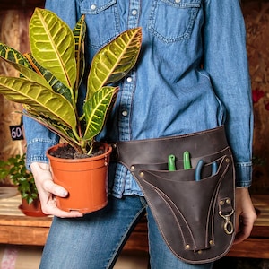 May include: A person wearing a blue denim shirt and jeans is holding a potted plant with green and yellow leaves. The person is wearing a brown leather tool belt with pockets and a metal clasp. The belt has gardening tools in the pockets.