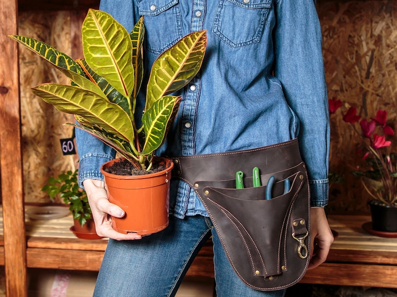 May include: A person wearing a blue denim shirt and jeans is holding a potted plant with green and yellow leaves. The person is wearing a brown leather tool belt with pockets and a metal clasp. The belt has gardening tools in the pockets.