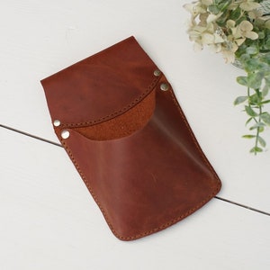 May include: A brown leather phone case with a flap and a snap closure. The case has a pocket on the front and is stitched around the edges.