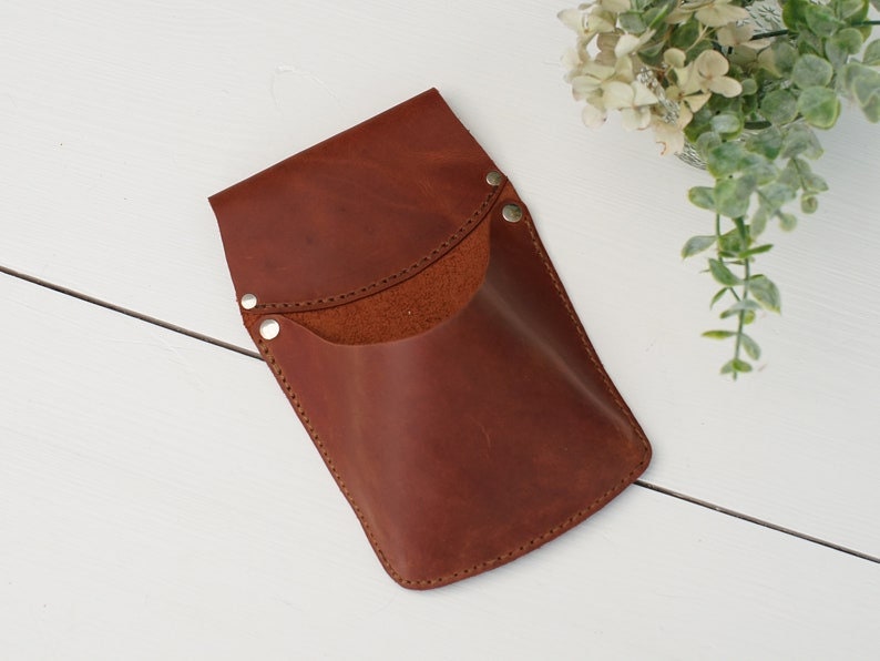 May include: A brown leather phone case with a flap and a snap closure. The case has a pocket on the front and is stitched around the edges.