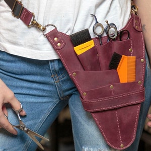 K&ouml;nnte beinhalten: Ein Werkzeugg&uuml;rtel aus burgunderfarbenem Leder mit mehreren Taschen und Riemen. Der G&uuml;rtel wird um die Taille getragen und enth&auml;lt verschiedene Friseurwerkzeuge, darunter Scheren, K&auml;mme und B&uuml;rsten.