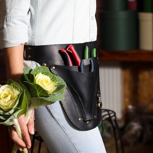 May include: A black leather gardening tool belt with a pouch and three gardening tools. The belt is worn around the waist of a person holding a bouquet of white cabbage roses.