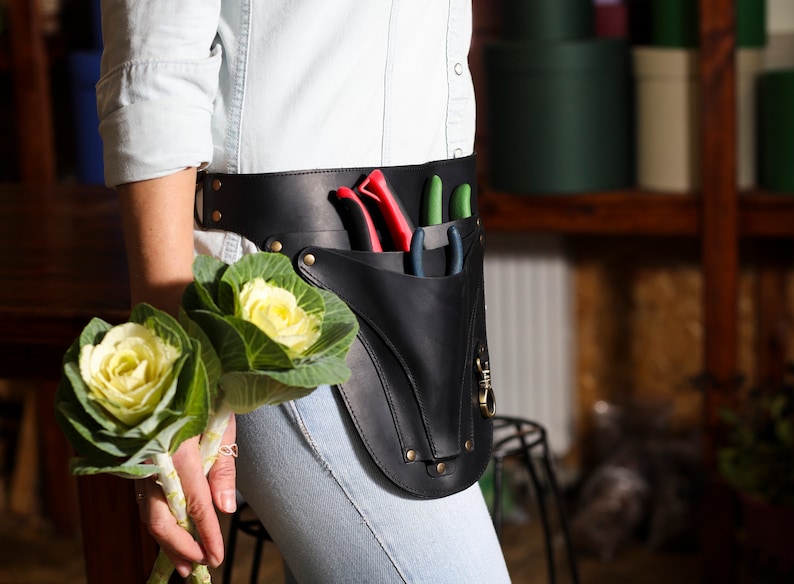 May include: A black leather gardening tool belt with a pouch and three gardening tools. The belt is worn around the waist of a person holding a bouquet of white cabbage roses.