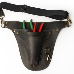 May include: A brown leather tool belt with a pocket for holding gardening shears. The belt has a black adjustable strap and a brass hook. Three shears are in the pocket, two green and one red.