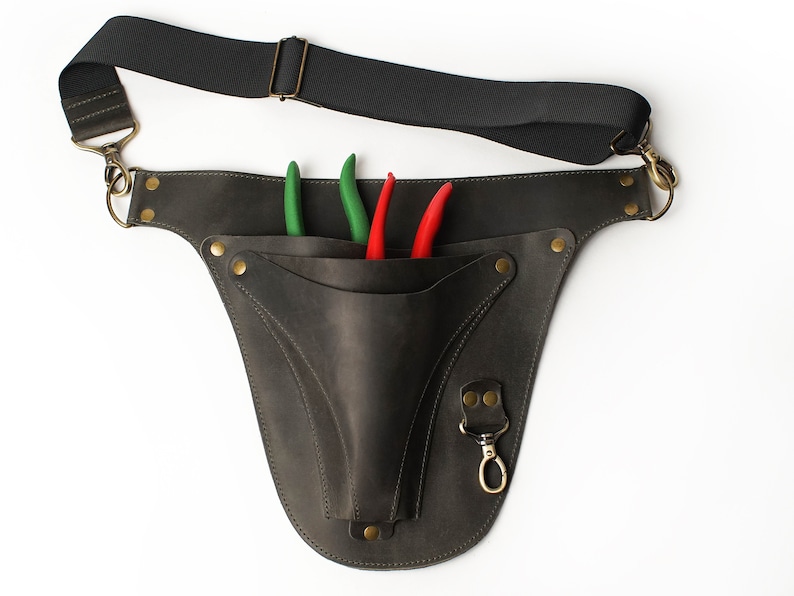 May include: A brown leather tool belt with a pocket for holding gardening shears. The belt has a black adjustable strap and a brass hook. Three shears are in the pocket, two green and one red.