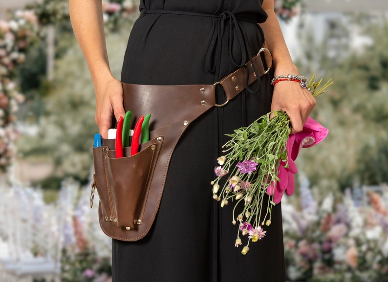 May include: A person wearing a black dress with a brown leather tool belt and pink gardening gloves. The belt has a pocket with gardening shears. The person is holding a bunch of flowers.