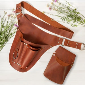 May include: A brown leather tool belt with multiple pockets and a detachable pouch. The belt has a wide strap and a metal buckle. The pouch has a single pocket and a metal ring.