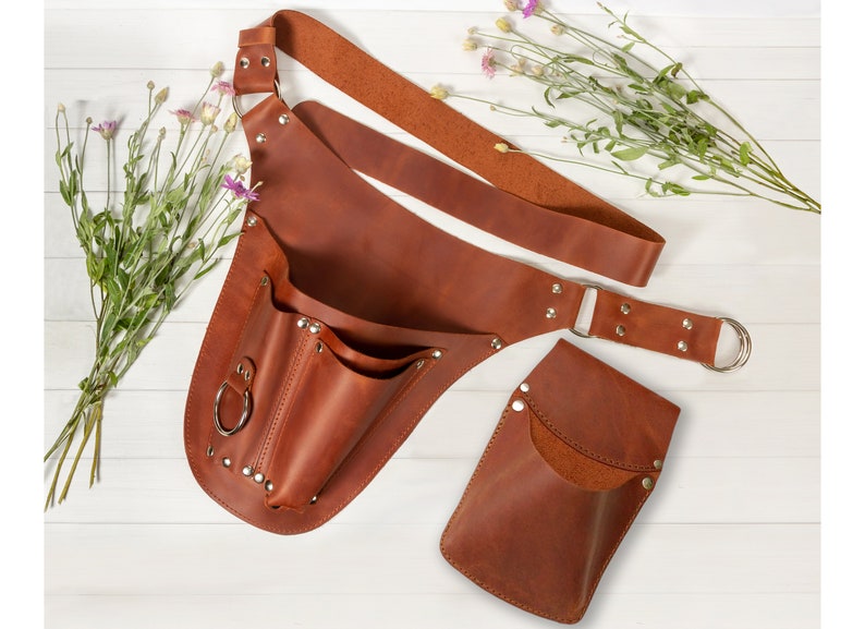 May include: A brown leather tool belt with multiple pockets and a detachable pouch. The belt has a wide strap and a metal buckle. The pouch has a single pocket and a metal ring.
