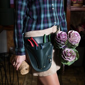 May include: A person wearing a green and blue plaid shirt and a brown leather belt with a green leather tool pouch. The pouch has several gardening tools, including pruning shears and a trowel. The person is holding a roll of twine in their hand. The person is standing in front of a wooden counter with a bouquet of purple ornamental cabbage.