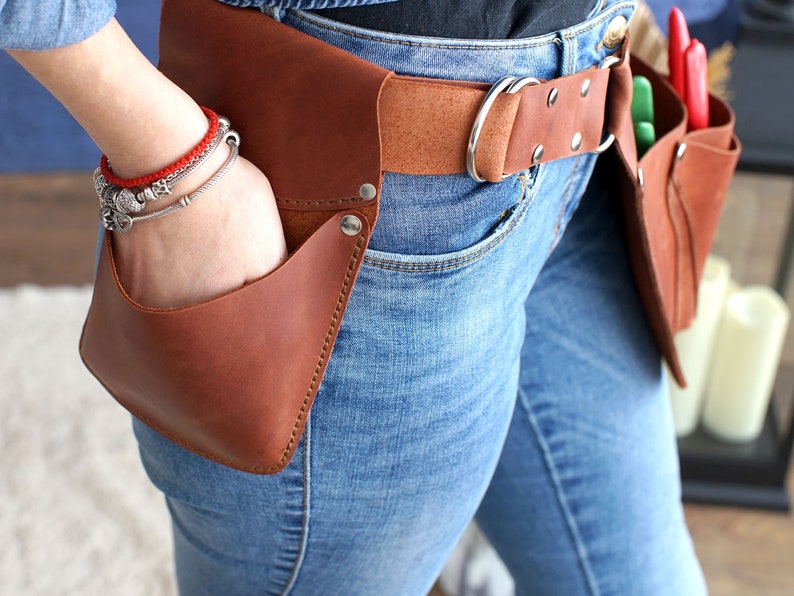 May include: A brown leather tool belt with multiple pockets and a brown leather belt with a silver buckle. The belt is worn over blue jeans.