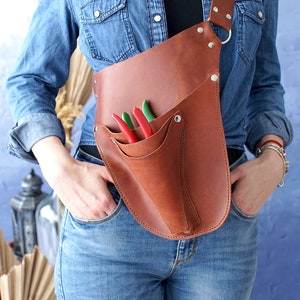 May include: A brown leather tool belt with a strap that goes over the shoulder. The belt has a pocket for tools and a sheath for a pair of pruning shears. The shears have red and green handles.