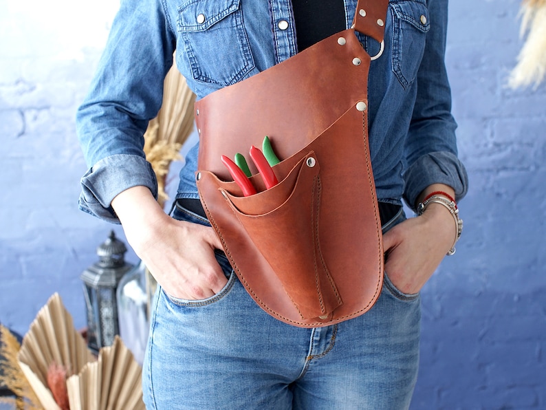 May include: A brown leather tool belt with a strap that goes over the shoulder. The belt has a pocket for tools and a sheath for a pair of pruning shears. The shears have red and green handles.