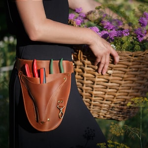 May include: A brown leather gardening tool belt with a pocket for holding shears and other tools. The belt is worn by a person holding a wicker basket filled with flowers.
