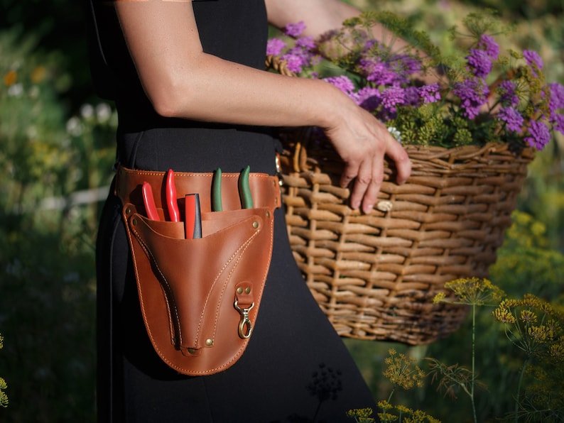 May include: A brown leather gardening tool belt with a pocket for holding shears and other tools. The belt is worn by a person holding a wicker basket filled with flowers.