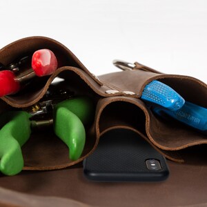 May include: A brown leather tool belt with multiple pockets, holding a pair of red pruning shears, a pair of green pruning shears, a pair of blue pruning shears, and a black phone.