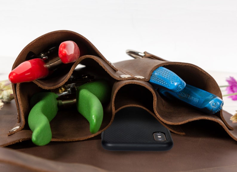 May include: A brown leather tool belt with multiple pockets, holding a pair of red pruning shears, a pair of green pruning shears, a pair of blue pruning shears, and a black phone.
