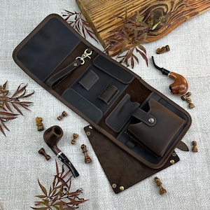 May include: A brown leather pipe case with multiple pockets and a snap closure. The case is open, revealing compartments for pipes and accessories. Two wooden pipes and chess pieces are also visible.