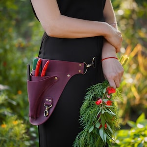 May include: A burgundy leather tool belt with a variety of gardening tools and a red strap. The belt is worn by a person holding a bouquet of flowers.