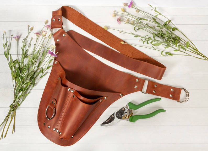 May include: A brown leather gardening tool belt with a large pocket and a smaller pocket. The belt has a metal buckle and a metal ring. A pair of green gardening shears are lying on the white surface next to the belt.