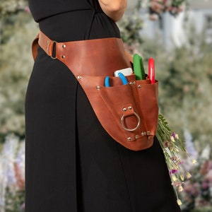 May include: A brown leather tool belt with multiple pockets and a metal ring. The belt is worn by a person and has various gardening tools in the pockets, including shears and a small knife.