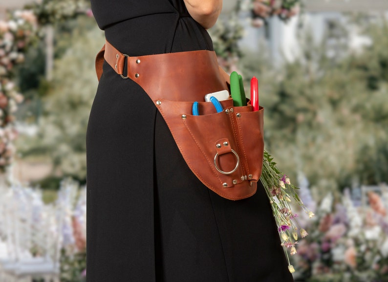 May include: A brown leather tool belt with multiple pockets and a metal ring. The belt is worn by a person and has various gardening tools in the pockets, including shears and a small knife.