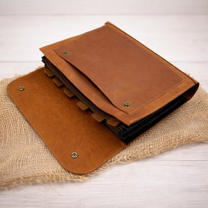 May include: Open, brown leather photo album with a rectangular design. The album has a flap closure with metal snap closures. The pages are black, and the album is resting on a burlap surface.