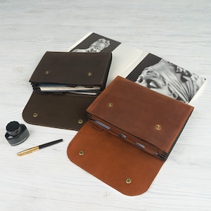 May include: Two brown leather document holders, one open and one closed, with brass snap closures. A black ink bottle and a gold-tipped pen are next to the document holders. A book with a black and white image is in the background.