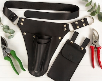 Könnte beinhalten: Schwarzer Ledergürtel für Gartengeräte mit zwei Taschen und einer abnehmbaren Tasche. Der Gürtel hat eine Schnalle und eine Schlaufe zum Befestigen an der Taille einer Person. Die Tasche hat eine Klappe mit einem Druckknopfverschluss. Der Gürtel ist auf einer weißen Oberfläche mit zwei Paar Gartenscheren abgebildet, eine grün und eine rot.