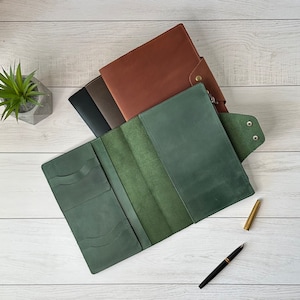 May include: A green leather notebook cover with multiple pockets and a flap closure. The cover is open to reveal a green interior with a brown leather notebook underneath.