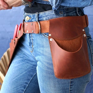 May include: A brown leather tool belt with multiple pockets and a metal buckle. The belt is worn over blue jeans.