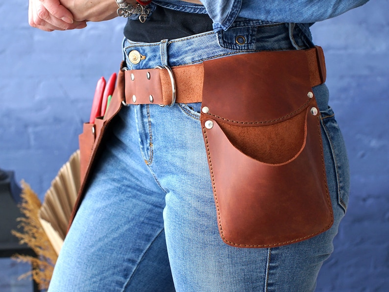May include: A brown leather tool belt with multiple pockets and a metal buckle. The belt is worn over blue jeans.