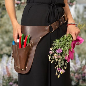 Puede incluir: Una persona lleva un vestido negro con un cinturón de herramientas de cuero marrón y guantes de jardinería rosas. El cinturón tiene un bolsillo con tijeras de jardín. La persona sostiene un ramo de flores.