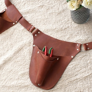 May include: A brown leather gardening tool belt with a large pocket and a smaller pocket on the side. The belt has a metal buckle and is secured with rivets. There are two green gardening shears in the pocket.