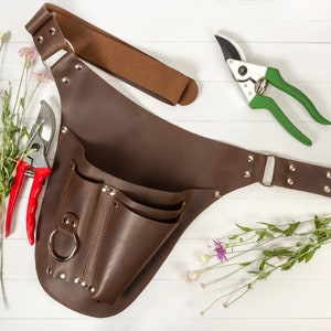 May include: A brown leather gardening tool belt with multiple pockets and a metal ring. The belt has a strap that can be adjusted to fit different sizes. The belt is shown with two pairs of gardening shears, one red and one green.