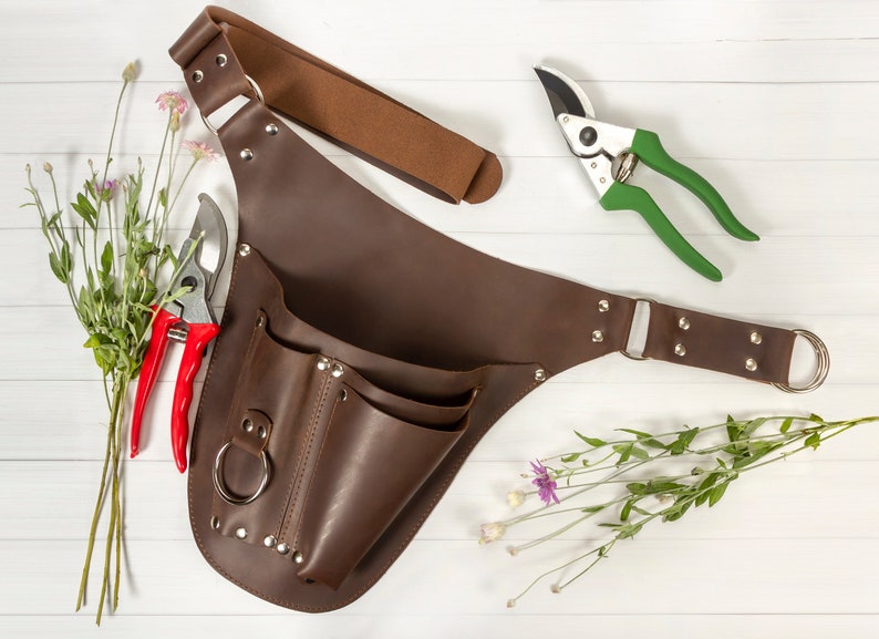 May include: A brown leather gardening tool belt with multiple pockets and a metal ring. The belt has a strap that can be adjusted to fit different sizes. The belt is shown with two pairs of gardening shears, one red and one green.