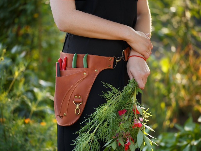 May include: A brown leather gardening tool belt with a variety of tools and a red string attached to the belt loop. The belt is worn by a person holding a bunch of green plants with red flowers.