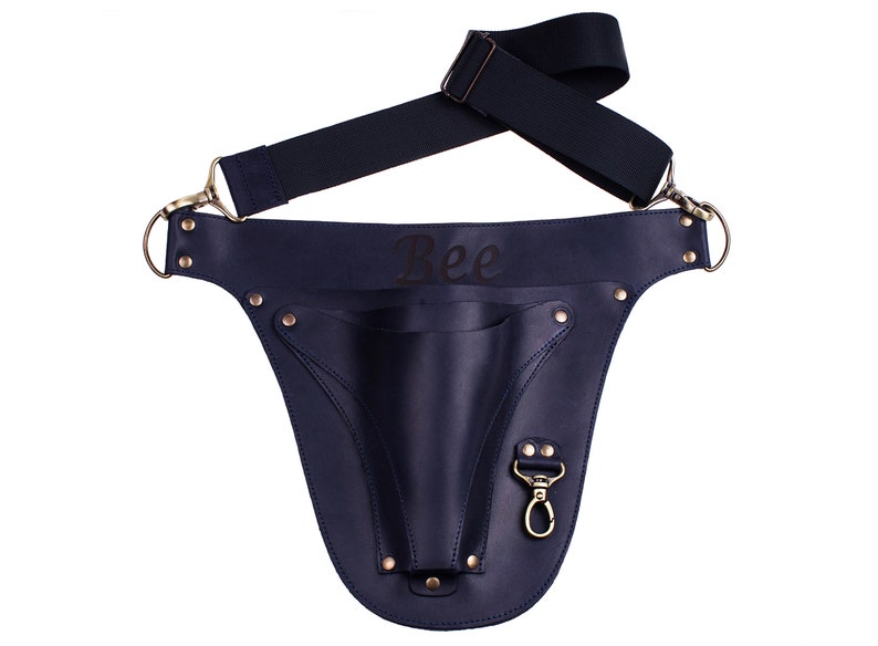 May include: A navy blue leather tool belt with a single pocket and a snap hook. The belt has a black adjustable strap and the word "Bee" is engraved on the front.