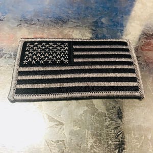 May include: A small, gray and black embroidered American flag patch. The flag is sewn onto a black fabric backing.