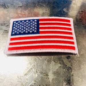 May include: A small, white-bordered patch with an embroidered American flag. The flag is red, white, and blue with 13 red and white stripes and a blue field of 50 white stars.